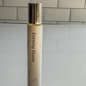 May include: A cylindrical perfume bottle with a gold cap. The label reads "Evening Bloom" with the name "Pamela Zinch" below. The bottle is filled with a light yellow liquid.