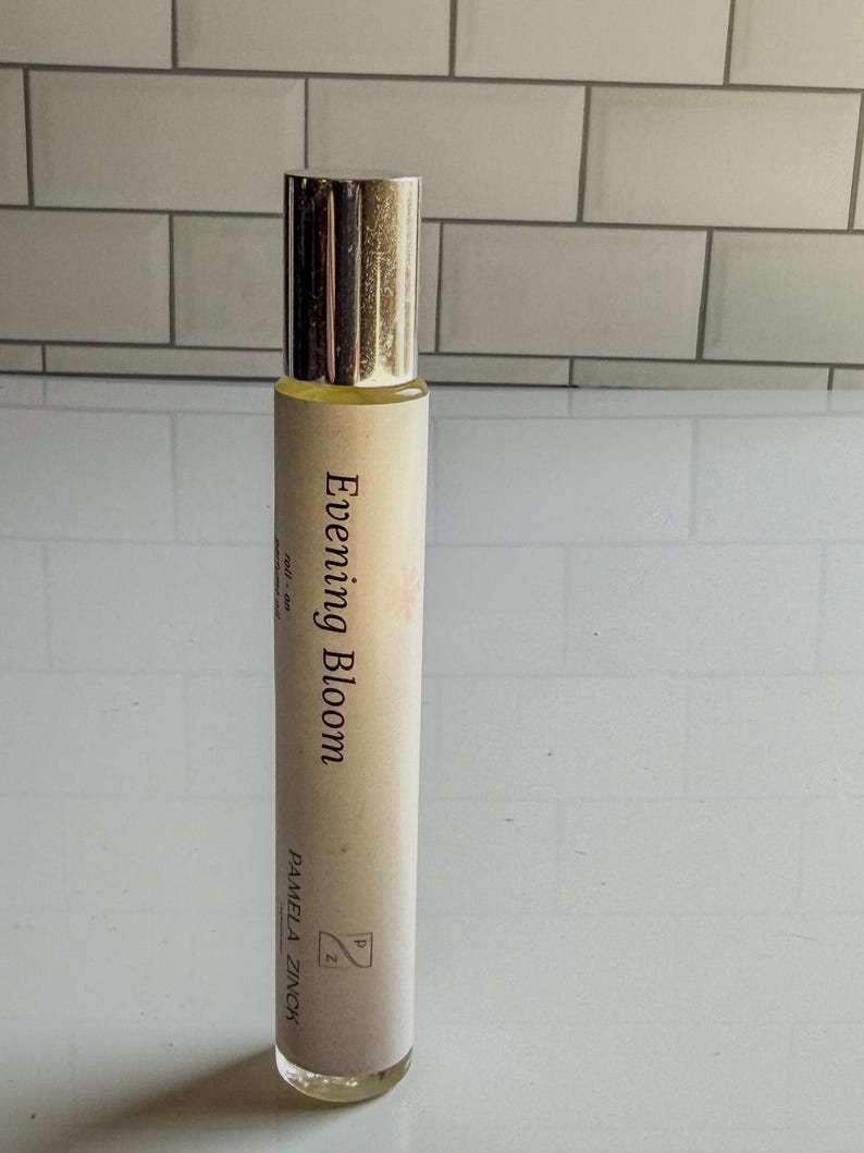 May include: A cylindrical perfume bottle with a gold cap. The label reads "Evening Bloom" with the name "Pamela Zinch" below. The bottle is filled with a light yellow liquid.