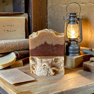 May include: Two bars of handmade soap, one brown and one beige, with a decorative swirl pattern. The soap is sitting on a wooden table next to an antique oil lamp.