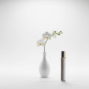 May include: A white ceramic vase holds a sprig of white orchids. A small perfume bottle with a gold cap and the text "Evening Bloom" is next to the vase. The background is white.