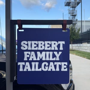 Puede incluir: Un letrero cuadrado de color azul oscuro con texto blanco que dice "SIEBERT FAMILY TAILGATE" en una fuente negrita y redondeada. El letrero está suspendido de un soporte de metal negro mediante dos correas marrones, frente a un vehículo negro.