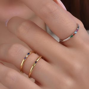 Peut inclure: Trois bagues sont présentées sur une main. Deux sont en or, chacune avec de petites pierres précieuses colorées. La troisième bague est en argent, également avec de petites pierres précieuses colorées. Les bagues sont délicates et élégantes.