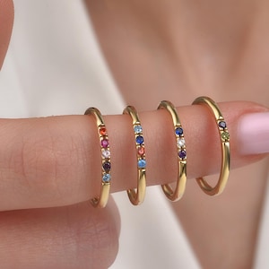 May include: Four gold rings, each featuring a line of colourful gemstones. The gemstones include red, pink, blue, purple, and green. The rings are displayed on a finger, highlighting their design and colour variations.