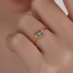 May include: A gold ring with two rectangular gemstones: a green and a purple one. The ring has a simple design, with the gemstones set next to each other on a thin band. The ring is shown on a finger.