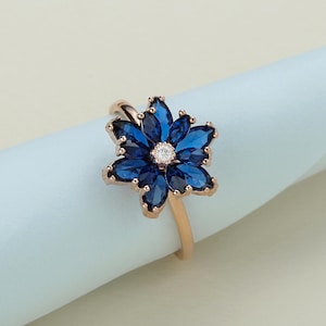 Peut inclure: Une bague en or rose avec un motif de fleur bleue. La fleur est composée de petites pierres bleues en forme de marquise avec une seule pierre ronde transparente au centre.