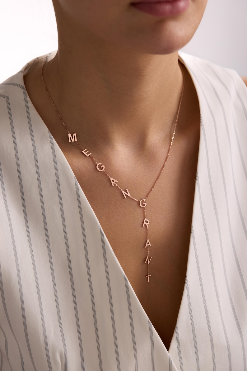May include: A rose gold necklace with the name "MEGAN GRANT" spelled out in individual letters. The necklace is worn around the neck and hangs down the chest. The letters are connected by a delicate chain.