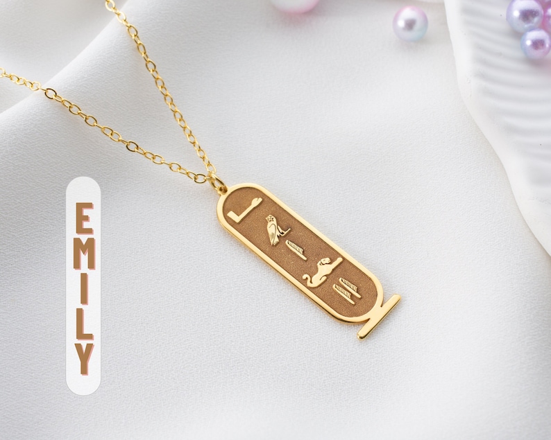 May include: Gold chain necklace with a pendant featuring an Egyptian hieroglyphic design. The pendant is rectangular and has a brown background with gold accents. The pendant features a cat, a bird, and other hieroglyphs.
