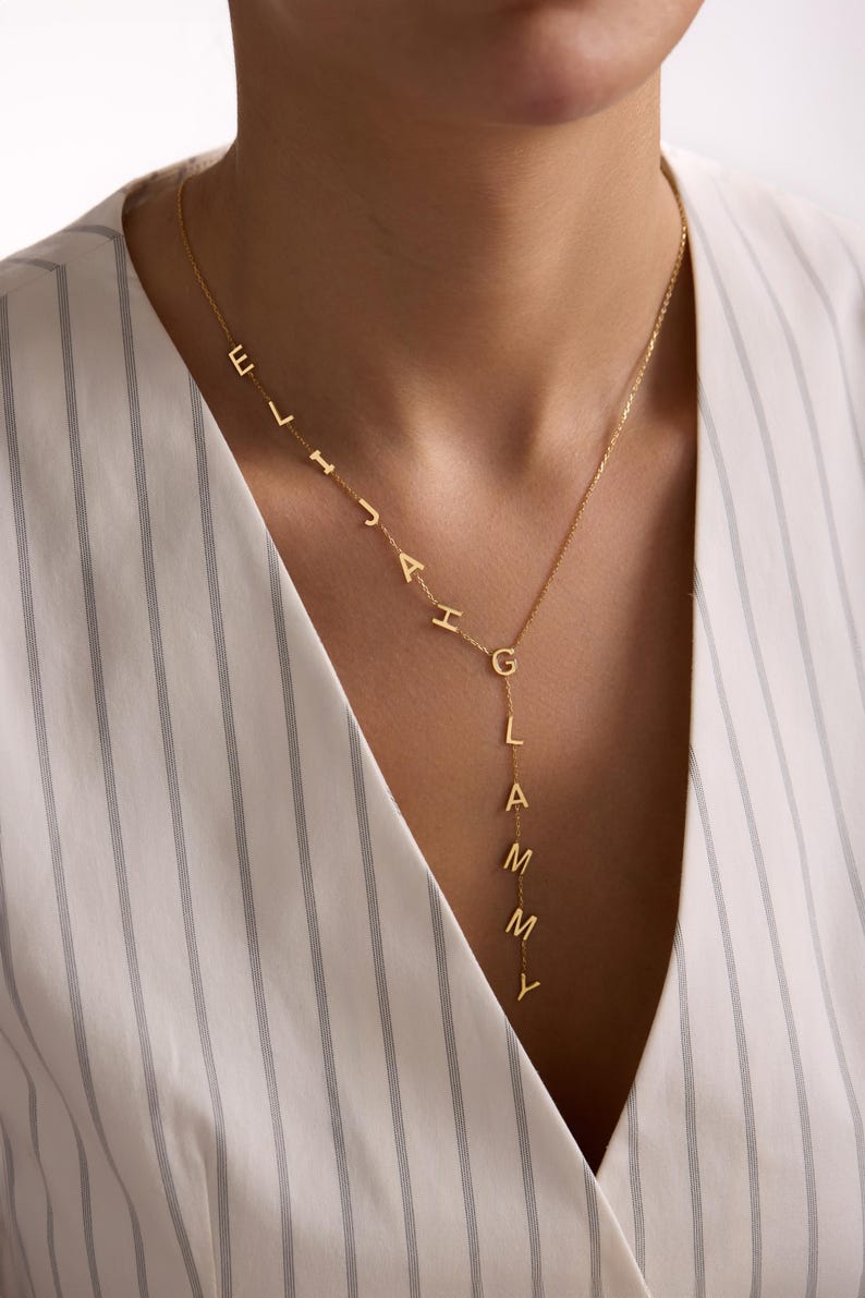 May include: A gold-colored necklace with the name "ELIJAH GLAMMY" spelled out in individual capital letters. The letters are connected by a delicate chain, creating a personalized and elegant accessory. The necklace is worn against a striped white and gray background.