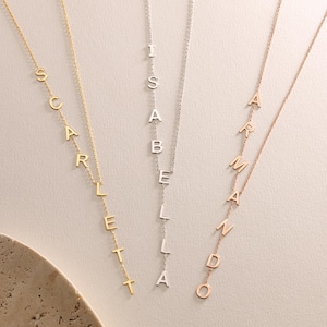May include: Three delicate necklaces with names spelled out in individual capital letters. The necklaces are in gold, silver, and rose gold. The names are Scarlett, Isabella, and Armando. The necklaces hang vertically.