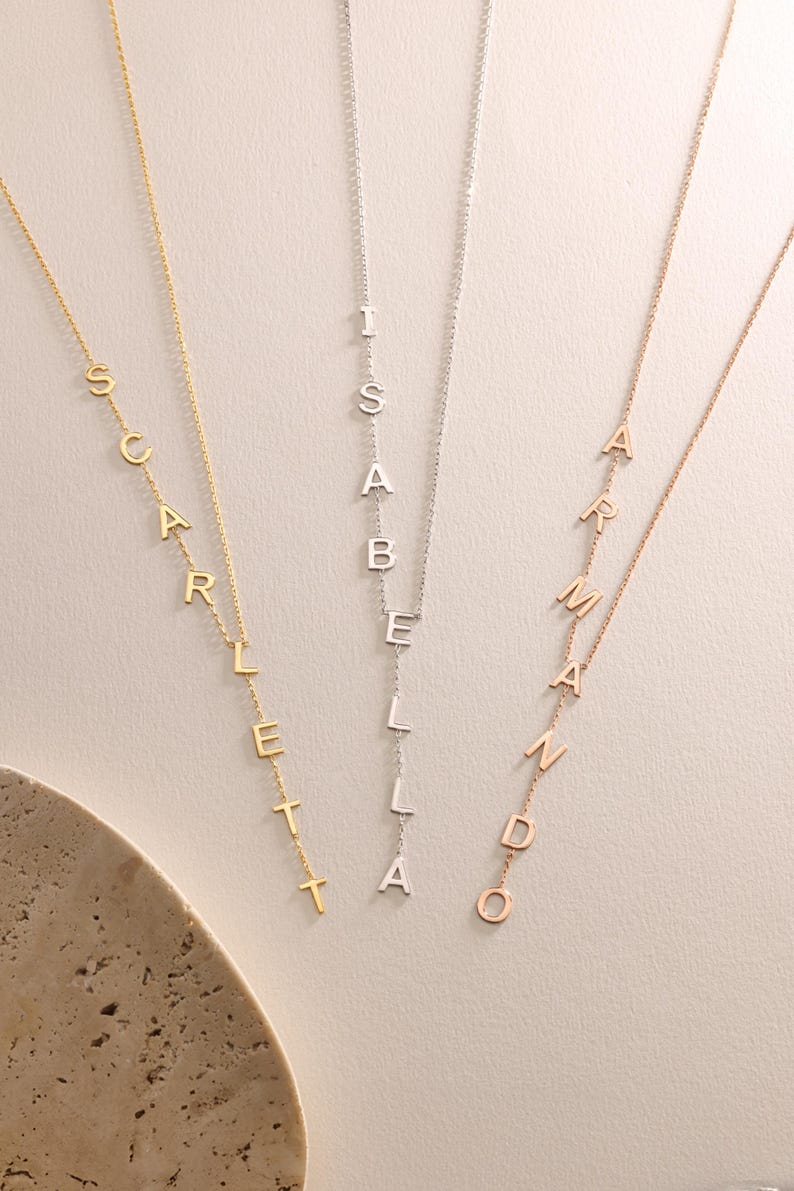 May include: Three delicate necklaces with names spelled out in individual capital letters. The necklaces are in gold, silver, and rose gold. The names are Scarlett, Isabella, and Armando. The necklaces hang vertically.