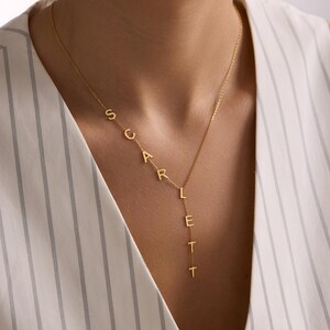 May include: A gold-colored necklace spelling out the name "SCARLETT" in individual capital letters. The necklace is worn around the neck and hangs down the chest. The background is a white and gray striped top.