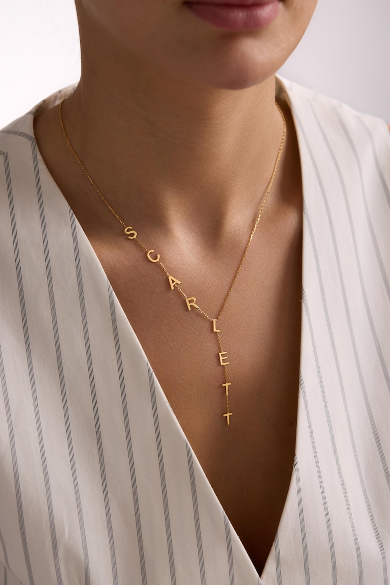 May include: A gold-colored necklace spelling out the name "SCARLETT" in individual capital letters. The necklace is worn around the neck and hangs down the chest. The background is a white and gray striped top.