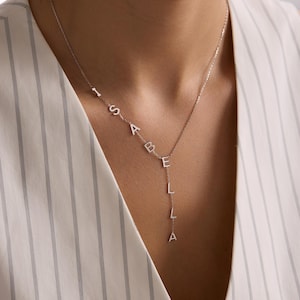 May include: A silver-colored necklace spelling out the name "ISABELLA" in individual capital letters. The letters are connected by a delicate chain, with the name cascading down the chest. The necklace is worn against a striped white and gray background.