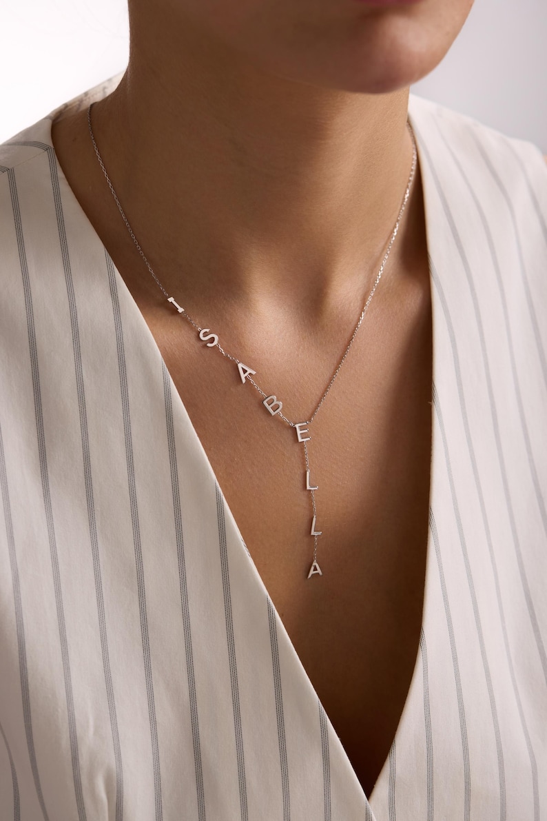 May include: A silver-colored necklace spelling out the name "ISABELLA" in individual capital letters. The letters are connected by a delicate chain, with the name cascading down the chest. The necklace is worn against a striped white and gray background.