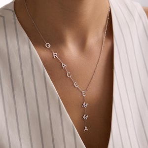 May include: A silver necklace spelling out the name "GRACE EMMA" in individual capital letters. The necklace is worn by a person wearing a white striped top. The letters are connected by a delicate chain.