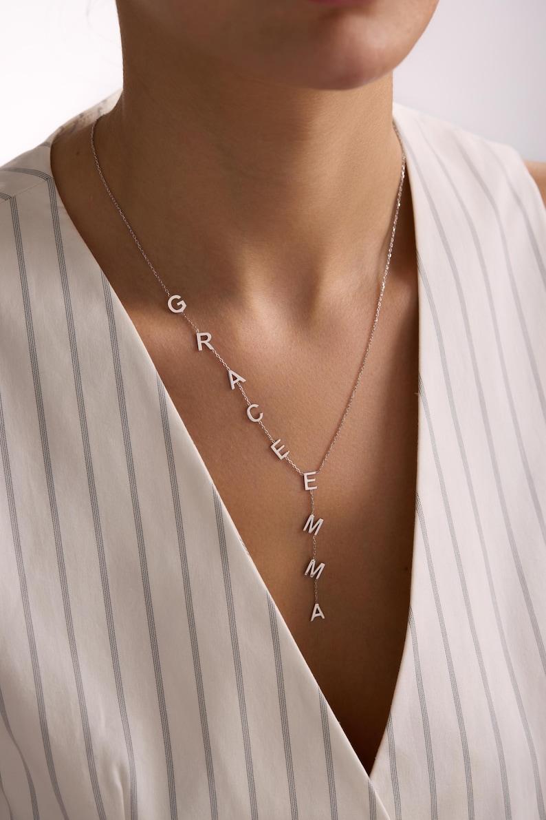 May include: A silver necklace spelling out the name "GRACE EMMA" in individual capital letters. The necklace is worn by a person wearing a white striped top. The letters are connected by a delicate chain.