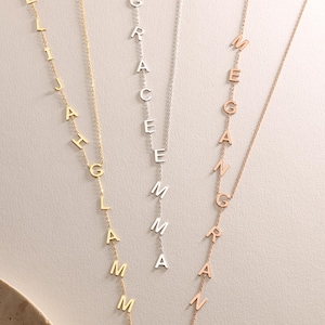 May include: Three personalized name necklaces in gold, silver, and rose gold. Each necklace features a vertical arrangement of individual letters spelling out a name. The necklaces hang against a neutral background.