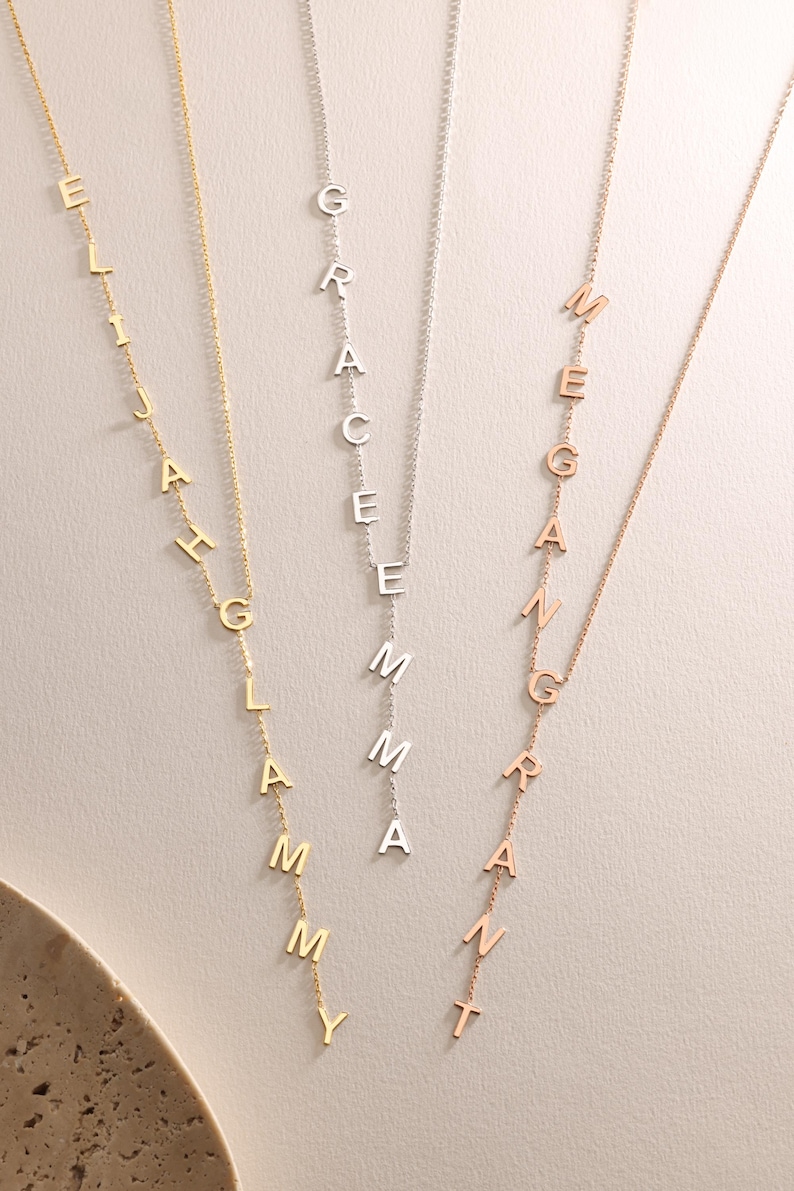 May include: Three personalized name necklaces in gold, silver, and rose gold. Each necklace features a vertical arrangement of individual letters spelling out a name. The necklaces hang against a neutral background.