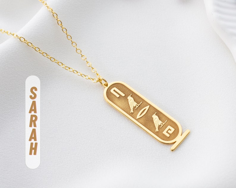 May include: Gold chain necklace with a pendant featuring the Egyptian hieroglyphs for the name "SARAH".
