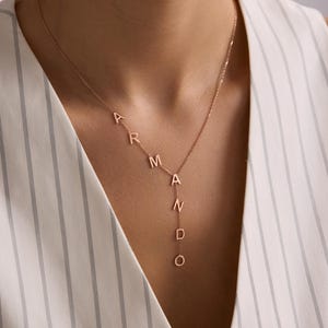 May include: Rose gold necklace spelling "Armando" in delicate, individual letters. The pendant hangs vertically, with the letters arranged in a stylish, personalized design. A simple and elegant piece of jewelry.