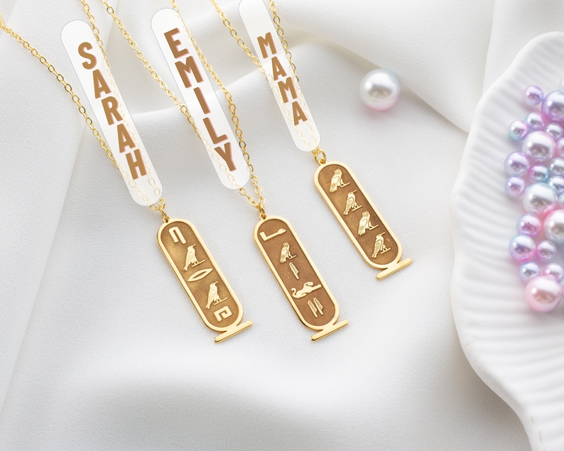 May include: Three gold necklaces with pendants featuring the Egyptian hieroglyphics for "Sarah", "Emily", and "Mama". Each pendant is a rectangular shape with a gold chain.