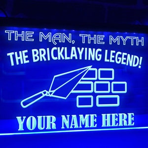 Puede incluir: Letrero LED azul con el texto "THE MAN, THE MYTH, THE BRICKLAYING LEGEND!" y "YOUR NAME HERE". El letrero presenta un diseño de llana y ladrillos. El letrero está iluminado con una luz azul brillante.