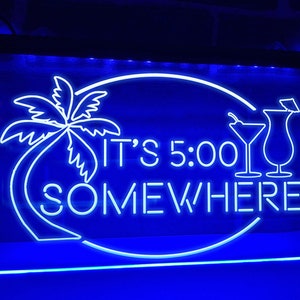 Op de afbeelding: Een rechthoekig blauw neonbord met de tekst "IT'S 5:00 SOMEWHERE" in het wit. Het bord toont een palmboom en cocktailglazen. Het bord wordt verlicht door een helder blauw licht.