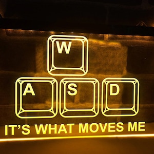 May include: Illuminated sign featuring the letters W, A, S, and D in a keyboard arrangement, with the text "IT'S WHAT MOVES ME" beneath. The sign emits a warm yellow glow, set against a brick wall.