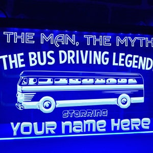 Könnte beinhalten: Blaues Neon-Schild mit weißem Text, der "The Man, The Myth, The Bus Driving Legend! Starring Your Name Here" lautet. Das Schild zeigt eine weiße Silhouette eines Oldtimerbusses.
