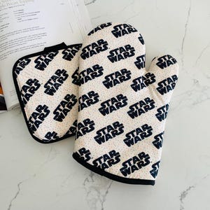 May include: A fabric oven mitt and a matching square pot holder featuring a repeating black Star Wars logo pattern on a cream-colored background with small multicolored speckles and black trim along the edges.