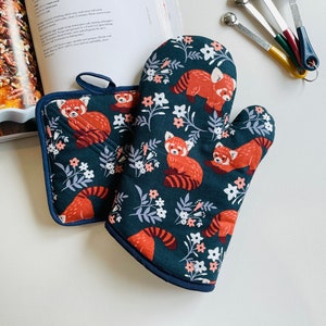 May include: A dark teal oven glove and pot stand set featuring a red panda and floral design. The oven glove and pot stand are trimmed with navy blue piping. The pot stand is square.