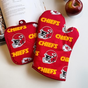 May include: A red oven mitt and a matching square pot holder featuring a repeating pattern of Kansas City Chiefs football helmets, the word CHIEFS in yellow text, and the team arrowhead logo. Both items have a quilted fabric construction.