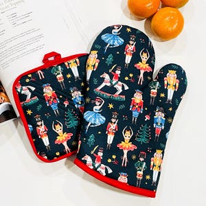 May include: A dark teal oven mitt and pot holder set with a red trim. The set features a whimsical print of nutcrackers, ballerinas, and Christmas trees. A cookbook and oranges are in the background.