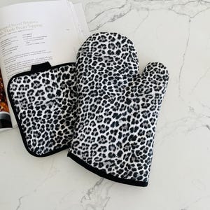 May include: A black and white leopard print oven mitt and pot holder set. The oven mitt is a standard size, and the pot holder is square. Both items have a black trim and are made of a soft, quilted material. A cookbook is open in the background.