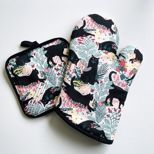 May include: A patterned oven mitt and pot holder set. The set has a white background with a repeating pattern of black cats, pink and yellow flowers, and green leaves. The edges are trimmed with black fabric.