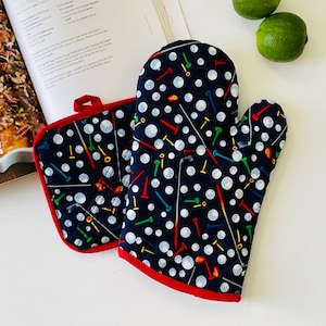 May include: A navy blue oven mitt and pot holder set with a red trim. The set features a pattern of white golf balls and colorful golf tees. The pot holder has a red loop for hanging. Kitchen and cooking accessories.
