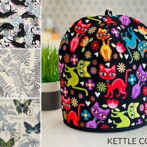 May include: A black kettle cozy with a colorful cat and flower pattern. The cozy is surrounded by fabric swatches featuring black cats, fern leaves, and butterfly designs. The text "KETTLE COZY" is at the bottom.