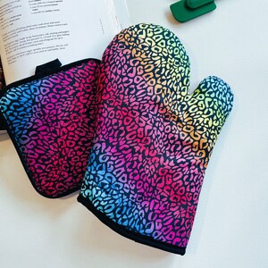May include: A colorful oven mitt and pot holder set. The set features a vibrant leopard print design in shades of pink, blue, yellow, and green. The edges are trimmed with black fabric. The items are designed for kitchen use.