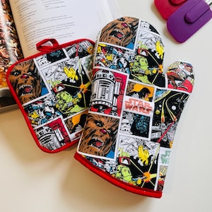 May include: A pair of red oven mitts with a Star Wars comic book print featuring characters like Chewbacca, R2-D2, and Darth Vader. The mitts are made of a thick, heat-resistant fabric.