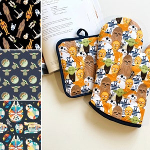 May include: A pair of Star Wars themed oven gloves. The gloves are orange and blue with a pattern of various Star Wars characters, including Yoda, Chewbacca, R2-D2, and C-3PO.