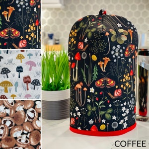 May include: A black coffee cozy with a red trim and a mushroom and floral pattern. The cozy is on a counter next to a French press.