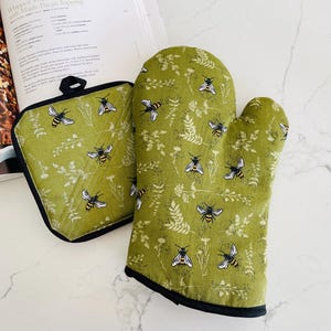 May include: A green oven mitt and pot holder set with a bee and floral pattern. The kitchen textiles have a black trim and a loop for hanging. The bee design is in black and yellow.