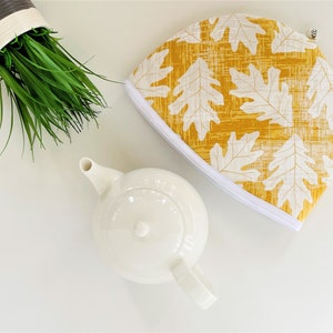 May include: A yellow tea cozy with a white oak leaf pattern. The cozy is made of fabric and has a white zipper around the edge.
