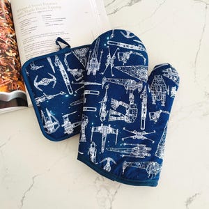 May include: A navy blue oven mitt and pot holder set. The set features a white schematic design of various space ships. The pot holder has a loop for hanging. The set is next to an open cookbook.