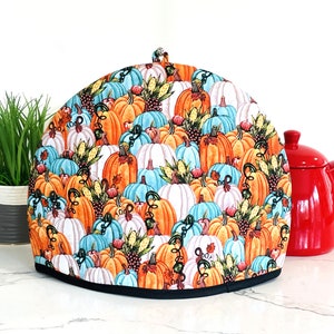 May include: A tea cozy with a colorful pumpkin pattern. The cozy features a black trim and a loop at the top. The background includes a red teapot and a potted plant, suggesting a fall or harvest theme.