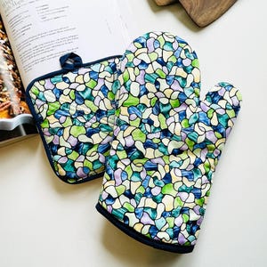 May include: A quilted oven mitt and pot holder set with a stained glass pattern in shades of blue, green, and purple. The set has a dark blue trim and loop. A cookbook is open in the background.