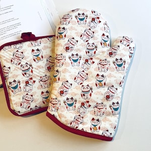 May include: A pair of red and white oven mitts with a pattern of lucky cats. The mitts are made of a soft, quilted fabric and have a red trim.