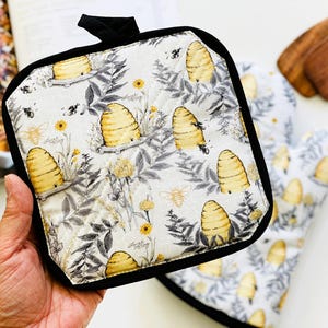 May include: A square quilted pot holder and a matching oven mitt featuring a repeating pattern of yellow beehives, bees, and gray botanical illustrations on a white background, finished with a solid black fabric trim and a hanging loop.