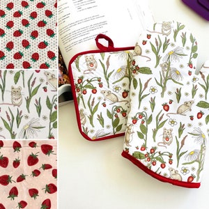 May include: Two red-trimmed oven mitts with a white background and a repeating pattern of mice, strawberries, and flowers. The mitts are on a white surface.