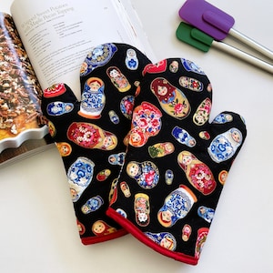 May include: Black oven mitts featuring a colourful pattern of Russian dolls, edged with red piping. The mitts are next to a cookbook and a dish of baked goods, with purple and green spatulas in the background.